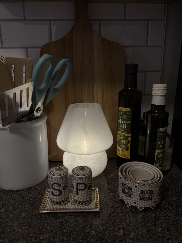 a moody kitchen counter with salt and pepper shakers, olive oil, a utensil jar and a small lit lamp
